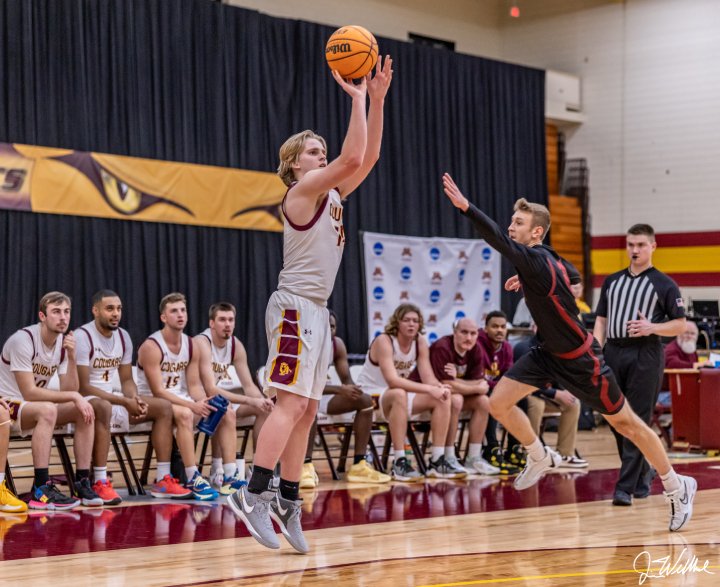 A basketball player in a white 'Cougars' jersey takes a jump shot while a defender in black leaps to block. Players on the bench watch in the background.